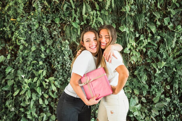 Happy female friends with pink gift box standing in front of green leaves