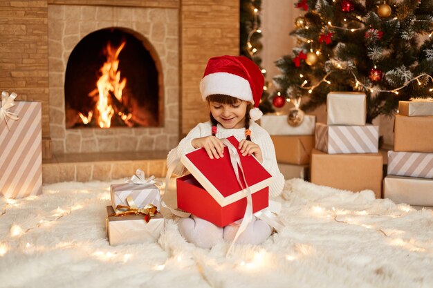 Happy female kid opening present box on New Year Eve, wearing white sweater and santa claus hat, sitting on floor near Christmas tree, present boxes and fireplace.