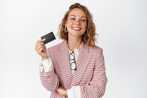 Happy female manager shows credit card and smiles pleased, stands in suit on white