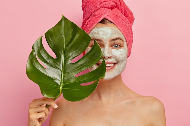 Happy female model has deep cleansing with facial mask, covers half of face with green leave, improves her appearance, wants to have fabulous skin, unclogs pores, smiles gently