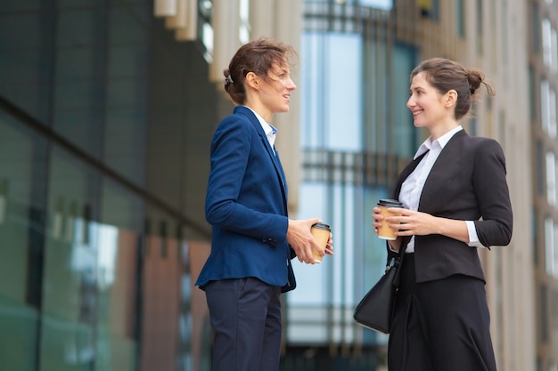 Happy female office friends with takeaway coffee cups meeting outdoors, talking, discussing project or chatting. Side view. Work break concept