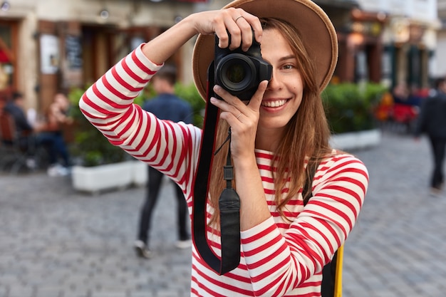 Happy female spends free time on hobby, takes picture of city street on camera during leisure