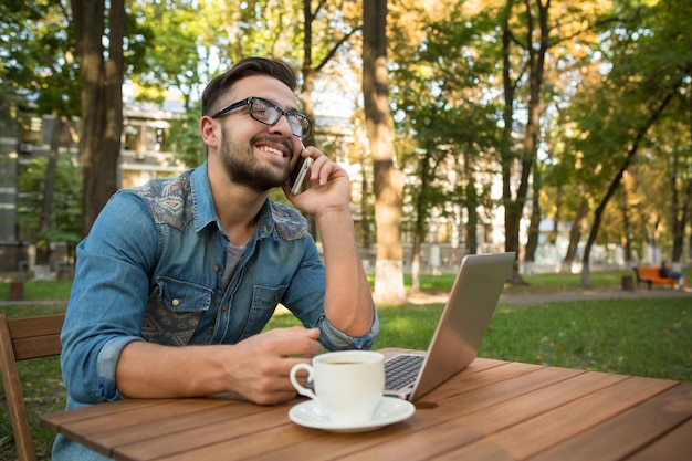 Happy freelancer hipster man speaking over mobile phone at the wooden table Smiling man using laptop computer and drinking coffee