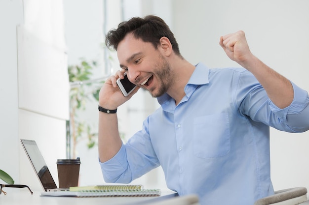 Free photo happy freelancer laughing while speaking over mobile phone cute man in blue shirt working on laptop computer indoors