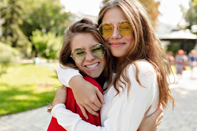 Happy friendly girls hugging each other with true smiles and having fun outside. Portrait  of two excited ladies expressing positive emotions enjoying walking.