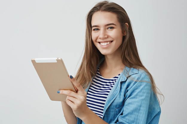 Happy friendly gorgeous female assistant smiling broadly holding digital tablet, posing joyfully, satisfied how easy draw using gadget