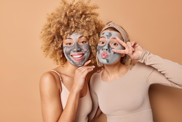 Free Photo happy friendly two women pose indoor with facial clay masks smile and make peace gesture over eye take care of skin and complexion dressed casually isolated over brown background beauty time