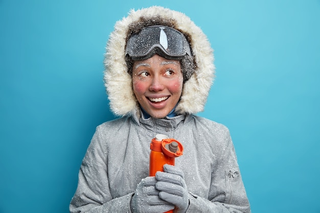 Free photo happy frosty woman in winter clothes warms during cold weather with hot tea from thermos smiles broadly wears ski goggles has active rest. brave positive female hiker.