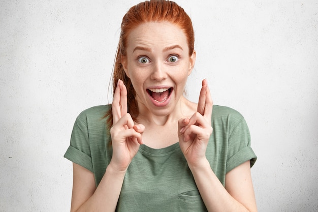 Free Photo happy funny overjoyed ginger female with hair knot, wears casual t shirt, crosses fingers with great hope, models in studio against white