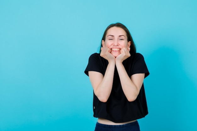 Happy girl is smiling by holding fists under chin on blue background