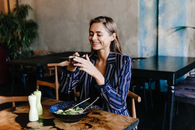 Free photo happy girl taking photo of her salad. indoor portrait of smiling brunette woman having fun during dinner.