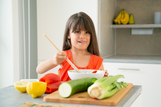 Free photo happy girl tossing salad in bowl with big wooden spoon. adorable kid learning to cook vegetables for dinner, posing, sticking out tongue. learning to cook concept