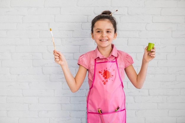 Free photo happy girl wearing pink apron holding paintbrush and paint bottle in hand