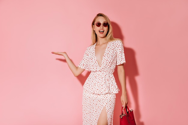 Free photo happy girl with blonde hair in red glasses and white dress holding cool handbag and pointing to place for text on pink background