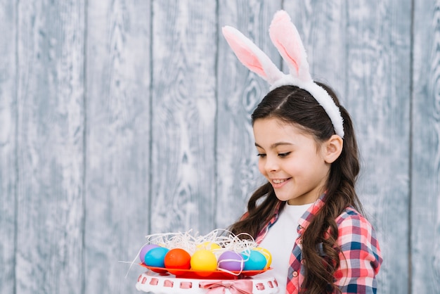 Happy girl with bunny ears looking at colorful easter eggs against wooden desk