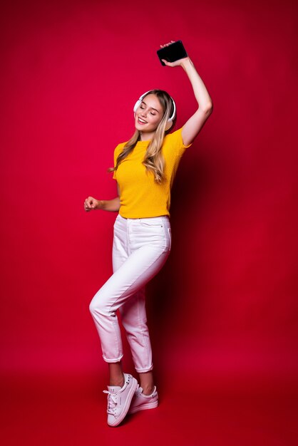 Happy girl with, listening to music in headphones and dancing, holding a smartphone. On a red wall.