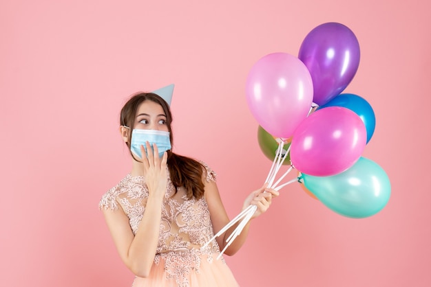 happy girl with party cap and medical mask holding colorful balloons on pink