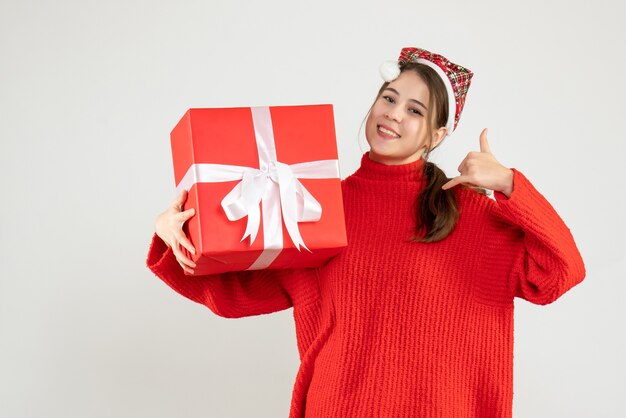 happy girl with santa hat holding present making call me sign standing on white