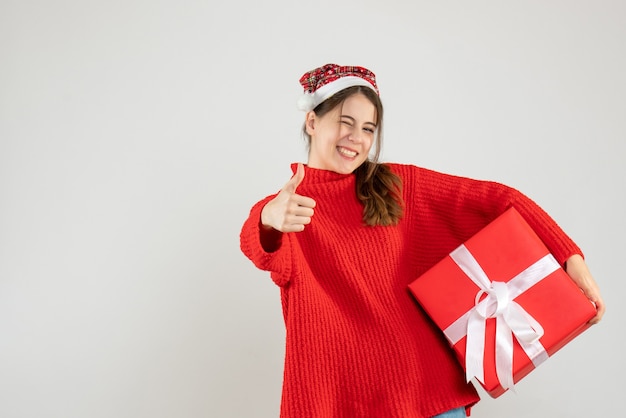 happy girl with santa hat making thumb up sign holding gift on white