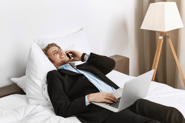 Happy good-looking manager with stylish haircut and beard lying on bed in hotel room, talking on phone and checking his work on laptop before business meeting