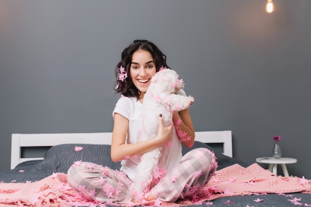 Happy good morning, true positive emotions of young joyful woman in pajamas with brunette curly hair having fun with little dog in pink tinsels on bed