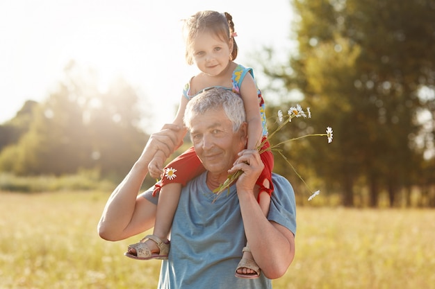 Happy grandfather and granddaughter have fun together outdoor. Grey haired male give piggyback ride to small child