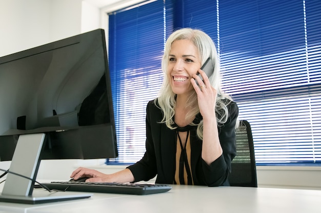 Free Photo happy grey-haired businesswoman talking with colleague on phone. professional confident manager chatting, smiling and working on computer. business, workplace and communication concept