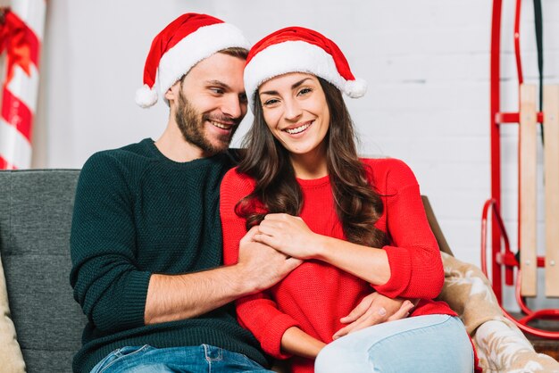 Happy guy and lady in party hats hugging on settee