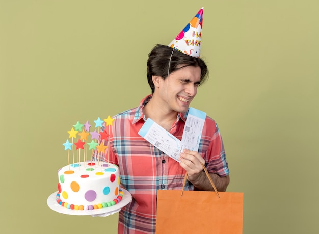 Happy handsome caucasian man wearing birthday cap holds paper shopping bag air tickets and birthday cake 