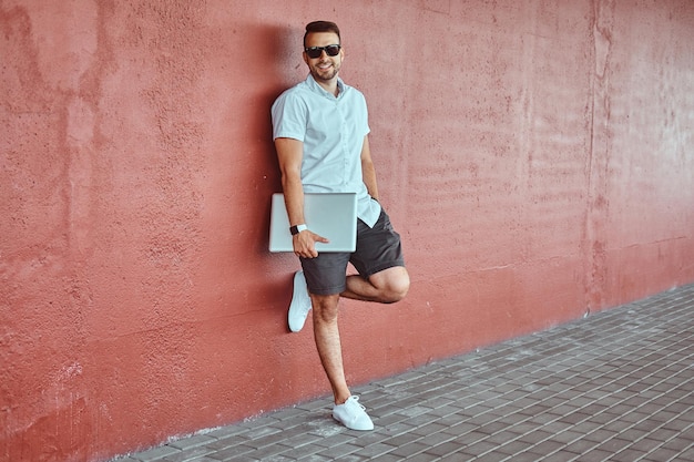 Free photo happy handsome fashionable freelancer in sunglasses dressed in a white shirt and shorts holds a laptop computer standing while leaning against a wall under a bridge.