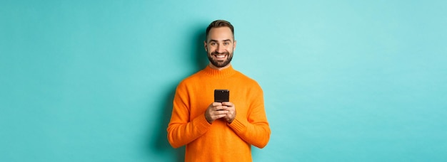 Free photo happy handsome man writing message on mobile phone holding smartphone and smiling standing against t