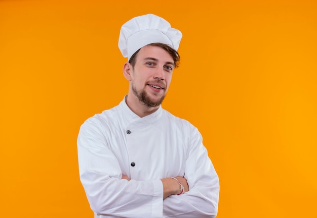 A happy handsome young bearded chef man in white uniform holding hands folded while looking on an orange wall