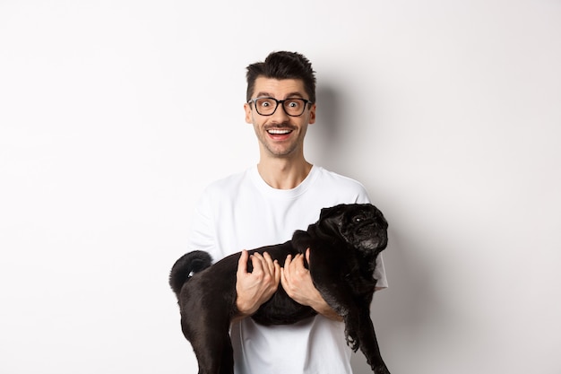 Free Photo happy hipster in glasses holding cute black pug and smiling, dog owner staring at camera with amazed smile, standing over white background.