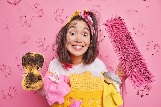 Free photo happy homemaker looks gladfully at you smiles toothily holds sponge and mop does spring cleaning of house cleans up apartment smeared with dirt poses near laundry basket isolated on pink