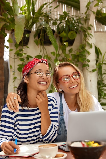 Happy interracial women embrace each other, sit in front of opened laptop, enjoy distance work from cafe