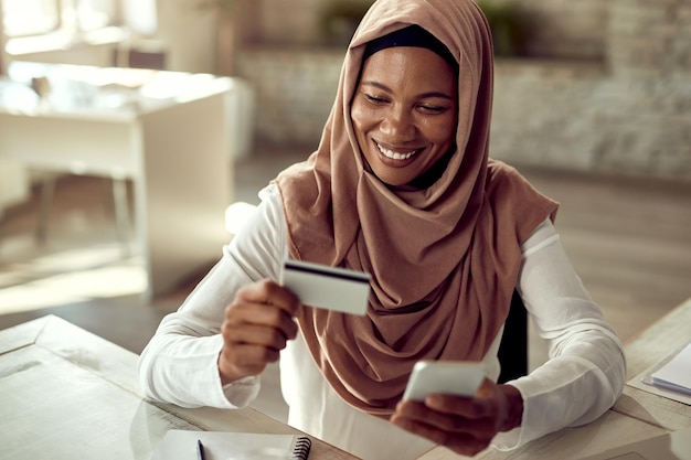 Happy Islamic businesswoman using credit card while online banking over smart phone in the office