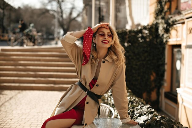 Happy joyful blonde curly woman in silk bright dress colorful sunglasses and beige trench coat sits on table and smiles outside