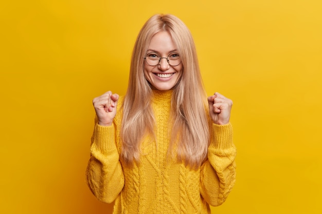 Happy joyful blonde woman clenches fists and celebrates good result smiles broadly being in high spirit makes triumph gesture wears spectacles and winter sweater 