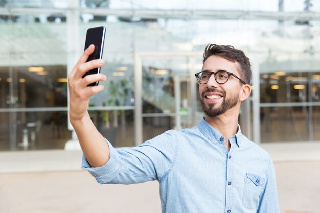 Happy joyful guy in eyewear taking selfie on smartphone