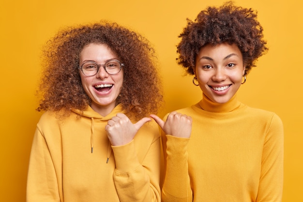 Happy joyful mixed race curly haired women point at each other with cheerful expression say its she stand closely to each other dressed casually isolated over yellow wall. I choose you