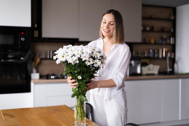 Happy and joyful young woman in white arranging white flowers at home in the kitchen