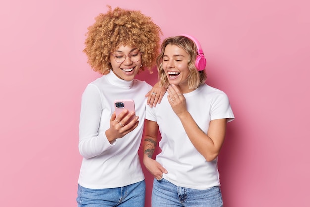 Happy joyful young women watch funny video on modern smartphone have fun listen music via headphones dressed in casual clothes pose against pink background People and modern technologies concept