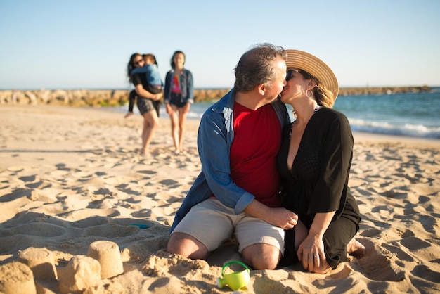 Happy kissing husband and wife at beach. Man and woman sitting at sand, hugging