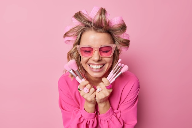 Happy lady applies hair rollers for making hairstyle wears trendy sunglasses sand blouse holds cosmetic brushes going to apply makeup smiles broadly isolated over pink wall Women beauty treatments