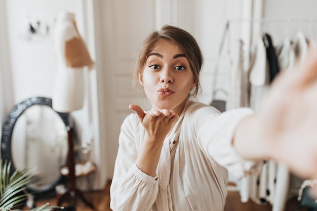 Free Photo happy lady in beige outfit blows kiss and takes selfie in office attractive woman in white blouse poses in good mood in apartment