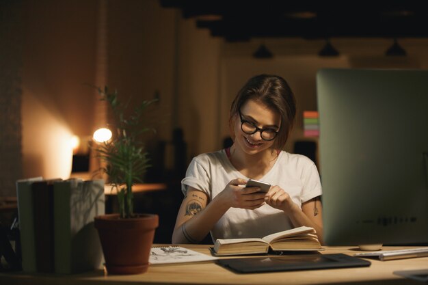 Happy lady designer sitting indoors at night chatting by phone.