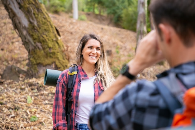 Free photo happy lady posing and smiling on road in forest. unrecognizable man taking photo of his girlfriend. tourists hiking together in woods and having fun. tourism, adventure and summer vacation concept