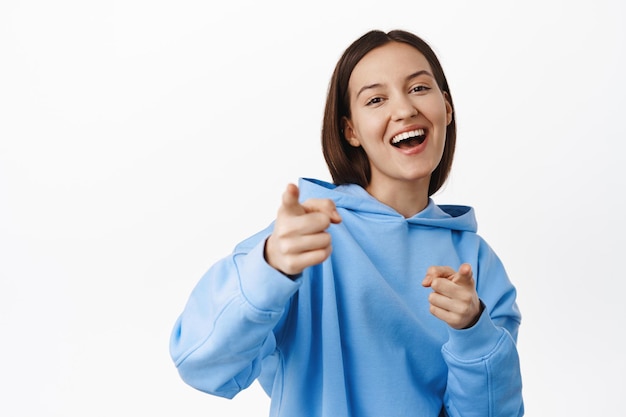 Happy laughing woman pointing fingers at camera and smiling, choosing you, congratulating, praising and picking person, standing in hoodie against white background