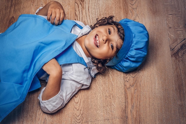 Free photo happy little boy with brown curly hair dressed in a blue cook uniform lying on a wooden floor.