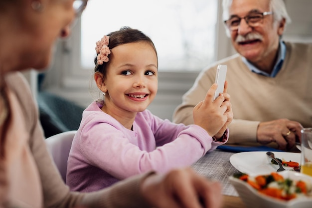 Happy little girl communicating with her grandparents while using cell phone during lunch at dining table